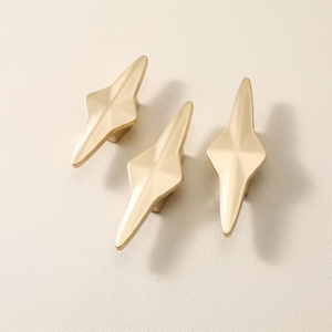 May include: Three gold-colored metal cabinet knobs with a pointed, elongated diamond shape. Each knob features a smooth, matte finish with faceted edges that taper to sharp points at the top and bottom, creating a symmetrical, geometric appearance.