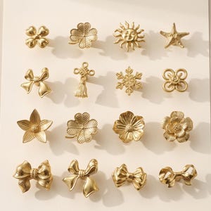 May include: A collection of sixteen gold-toned metal cabinet knobs arranged in a four-by-four grid. The designs include various shapes such as bows, flowers, a sun, a starfish, a snowflake, a four-leaf clover, and decorative knot motifs.