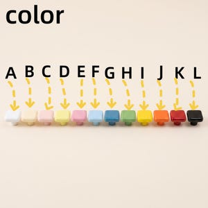 Puede incluir: Una fila de doce pomos cuadrados para armarios en varios colores, etiquetados de la A a la L. Los colores incluyen blanco, crema, rosa claro, amarillo p&aacute;lido, rosa, azul claro, azul, verde, amarillo, naranja, rojo y negro, con acabado brillante.