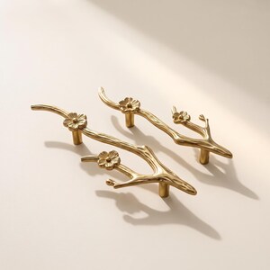 May include: Two gold-toned metal cabinet pulls shaped like tree branches with textured bark details and small, five-petaled flower accents. Each handle features two cylindrical mounting posts on the underside for attachment to furniture or cabinetry.