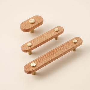 Puede incluir: Tres tiradores de armario oblongos de distintas longitudes, fabricados en madera de color marrón claro con veta visible. Cada tirador cuenta con postes de montaje metálicos circulares y tapas de tornillo planas y redondas.