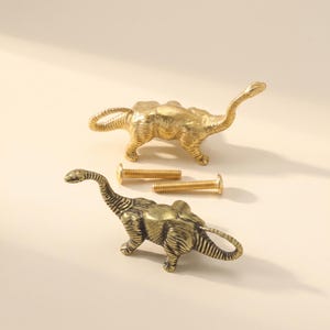 Pode incluir: Dois puxadores de armário em metal dourado com formato de dinossauros de pescoço longo. Cada puxador apresenta um corpo texturizado com uma cauda curva e um pescoço longo. Dois parafusos de metal dourado a condizer estão entre os puxadores.