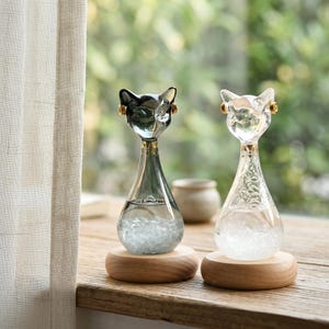 Pode incluir: Dois termômetros decorativos em forma de gato de vidro. Um é transparente, o outro cinza. Ambos têm uma base em forma de gota cheia de líquido e cristais, e assentam em bases redondas de madeira. As cabeças de gato têm detalhes dourados.