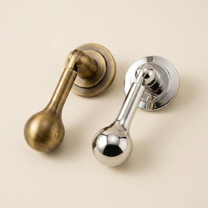 May include: Two pendant-style cabinet pulls, one with a brushed antique brass finish and the other with a polished chrome finish. Each pull features a circular mounting base and a teardrop-shaped handle that hangs from a hinged joint mechanism.