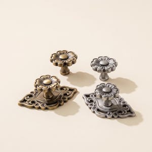 Può includere: Quattro pomelli per mobili in metallo con due finiture. Due pomelli presentano una testa semplice a forma di fiore, mentre gli altri due sono montati su placche decorative ornate con volute. Un set è in ottone anticato, l'altro in argento.