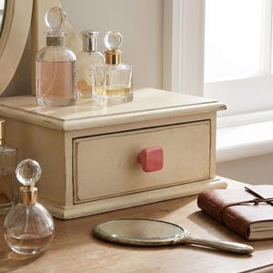 Puede incluir: Un organizador de tocador de madera color crema con un caj&oacute;n que tiene un pomo cuadrado rosa. Encima hay tres frascos de perfume de vidrio con tapones esf&eacute;ricos. Un espejo de mano ovalado y un diario de cuero marr&oacute;n est&aacute;n en primer plano.