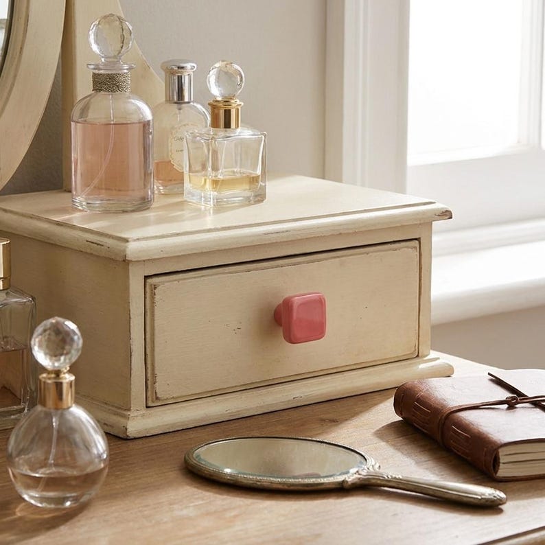 Puede incluir: Un organizador de tocador de madera color crema con un caj&oacute;n que tiene un pomo cuadrado rosa. Encima hay tres frascos de perfume de vidrio con tapones esf&eacute;ricos. Un espejo de mano ovalado y un diario de cuero marr&oacute;n est&aacute;n en primer plano.