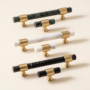 May include: A collection of six cabinet pulls and knobs featuring cylindrical stone bars in dark green, white, and black marble, each accented with brushed brass metal mounts. The set includes three long horizontal pulls and three smaller knobs.