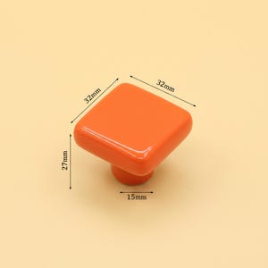Puede incluir: Un pomo de mueble cuadrado de color naranja con bordes redondeados. El pomo presenta l&iacute;neas de dimensi&oacute;n impresas que indican un ancho de 32 mm, un largo de 32 mm, una altura total de 27 mm y un di&aacute;metro de base de 15 mm. La superficie es lisa y brillante.