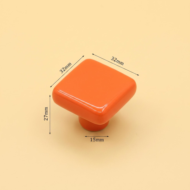 Puede incluir: Un pomo de mueble cuadrado de color naranja con bordes redondeados. El pomo presenta l&iacute;neas de dimensi&oacute;n impresas que indican un ancho de 32 mm, un largo de 32 mm, una altura total de 27 mm y un di&aacute;metro de base de 15 mm. La superficie es lisa y brillante.