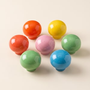 Op de afbeelding: Een verzameling van zeven bolvormige kastknoppen in verschillende glanzende kleuren, waaronder oranje, geel, roze, groen en blauw. Elke knop heeft een glad, afgerond bovenoppervlak en een kleine ronde basis voor montage op meubels.
