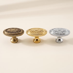 May include: Three round metal cabinet knobs featuring a decorative embossed floral pattern on the top surface and a beaded edge. The set includes one antique brass finish knob, one polished gold finish knob, and one polished chrome finish knob.