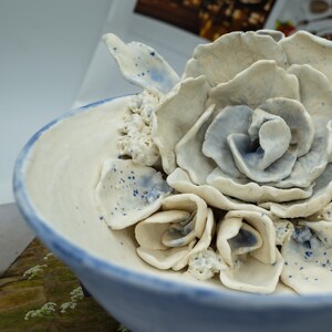 May include: A ceramic bowl with a floral design. The bowl is white with a blue rim and accents. The flowers are a mix of white and light blue, with a textured appearance. The bowl is likely handmade.