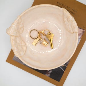 May include: A round, cream-coloured ceramic bowl with textured, leaf-like side handles. Inside the bowl sits a gold-toned keyring featuring a small, sparkling cup-shaped charm, a rectangular tag, and a standard metal house key.
