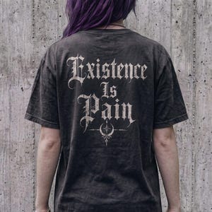 以下が含まれることがあります： 背面に掠れた白のゴシック体で「Existence Is Pain」という文字がプリントされた、チャコールグレーの半袖Tシャツ。文字の下には、三日月と星のデザインが施された小さく複雑な円形のシンボルが描かれています。