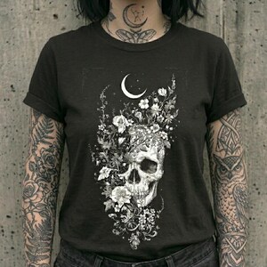 May include: A black short-sleeved t-shirt featuring a white graphic print of a human skull surrounded by various flowers and foliage, positioned beneath a crescent moon. The design is centered on the chest of the garment.