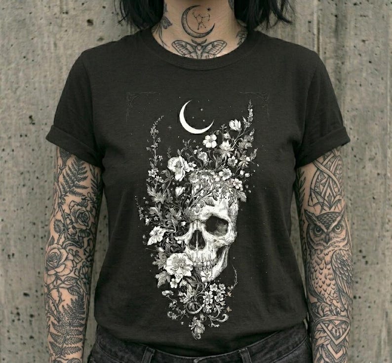 May include: A black short-sleeved t-shirt featuring a white graphic print of a human skull surrounded by various flowers and foliage, positioned beneath a crescent moon. The design is centered on the chest of the garment.