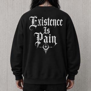 以下が含まれることがあります： 背面にゴシック体で「Existence Is Pain」と白文字でプリントされた黒の長袖スウェットシャツ。テキストの下には、星を伴う三日月と装飾的なフィリグリー模様が白で描かれています。