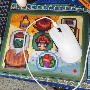 K&ouml;nnte beinhalten: Eine wei&szlig;e Computermaus mit Kabel liegt auf einem pixeligen Mauspads mit bunten Lebensmittel- und Charakterdesigns. Das Mauspad hat einen gr&uuml;nen Rand und liegt auf einer gemusterten Oberfl&auml;che.
