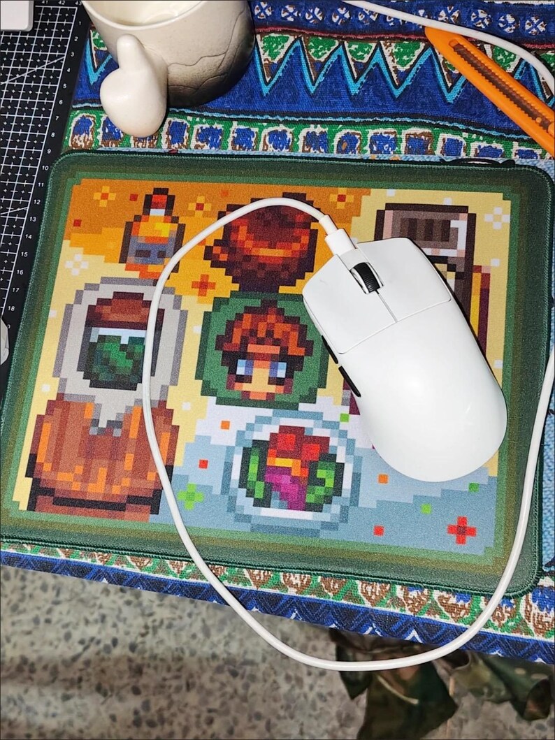 K&ouml;nnte beinhalten: Eine wei&szlig;e Computermaus mit Kabel liegt auf einem pixeligen Mauspads mit bunten Lebensmittel- und Charakterdesigns. Das Mauspad hat einen gr&uuml;nen Rand und liegt auf einer gemusterten Oberfl&auml;che.