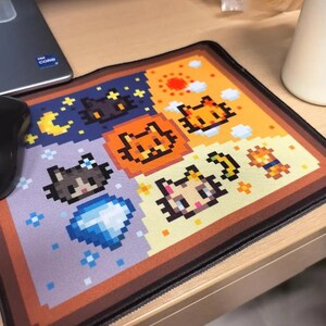 K&ouml;nnte beinhalten: Ein rechteckiges Mauspad mit pixeligen Katzendesigns in einem Rastermuster. Das Farbschema umfasst Orange, Gelb, Blau und Braun. Das Mauspad liegt auf einem Holztisch, mit einer Computermaus links.