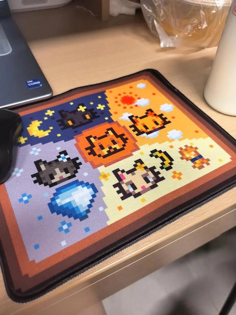 K&ouml;nnte beinhalten: Ein rechteckiges Mauspad mit pixeligen Katzendesigns in einem Rastermuster. Das Farbschema umfasst Orange, Gelb, Blau und Braun. Das Mauspad liegt auf einem Holztisch, mit einer Computermaus links.