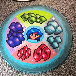 K&ouml;nnte beinhalten: Rundes Mauspad mit pixeligem Design von Edelsteinen, einem Charakter und anderen Spielelementen. Das Design umfasst gr&uuml;ne, blaue, rote und violette Edelsteine mit einem t&uuml;rkisfarbenen Rand. Das Mauspad ist f&uuml;r Spiele konzipiert.