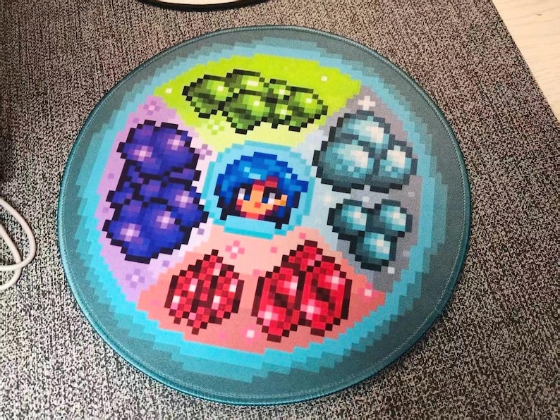 K&ouml;nnte beinhalten: Rundes Mauspad mit pixeligem Design von Edelsteinen, einem Charakter und anderen Spielelementen. Das Design umfasst gr&uuml;ne, blaue, rote und violette Edelsteine mit einem t&uuml;rkisfarbenen Rand. Das Mauspad ist f&uuml;r Spiele konzipiert.