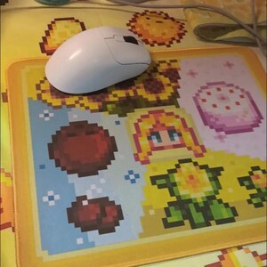 K&ouml;nnte beinhalten: Eine wei&szlig;e Computermaus liegt auf einem pixeligen Mauspad mit gelbem Rand. Das Mauspad zeigt farbenfrohe Pixelkunst eines Sonnenblumen, einer Figur und anderer spielbezogener Grafiken. Eine kleine geh&auml;kelte Sonnenblume steht in einem wei&szlig;en Topf.