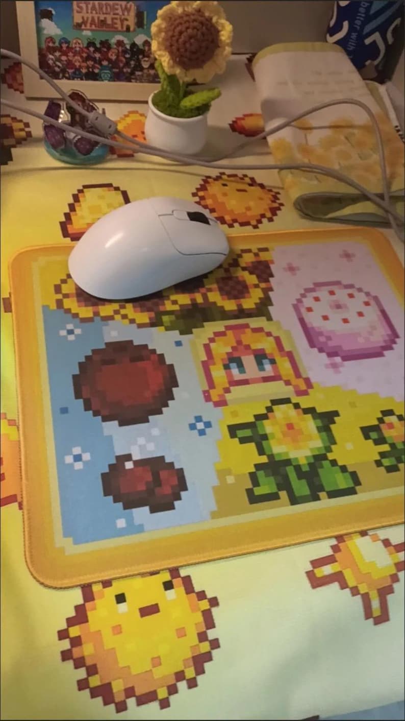 K&ouml;nnte beinhalten: Eine wei&szlig;e Computermaus liegt auf einem pixeligen Mauspad mit gelbem Rand. Das Mauspad zeigt farbenfrohe Pixelkunst eines Sonnenblumen, einer Figur und anderer spielbezogener Grafiken. Eine kleine geh&auml;kelte Sonnenblume steht in einem wei&szlig;en Topf.