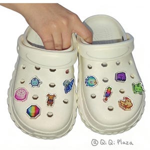 May include: A pair of white clogs adorned with colourful pixel art charms. The charms feature various shapes and characters. The clogs are being held by a hand. The text "@ Qi Qi Plaza" is visible.
