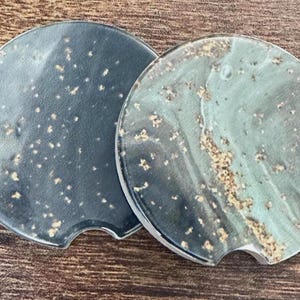 May include: Two round car coasters with a unique design. One coaster is dark blue with small white and gold speckles. The other is teal with gold and white marbling. Each coaster has a small notch for easy removal.