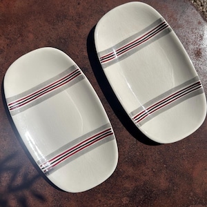 Può includere: Due piatti da portata ovali in ceramica bianco sporco. Ogni piatto ha due serie di strisce parallele grigie, rosse e nere. I piatti sono su una superficie scura e strutturata.