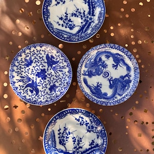 Puede incluir: Cuatro platos decorativos pequeños, redondos, blancos y azules. Cada plato presenta un diseño azul intrincado diferente, que incluye patrones florales, un dragón y un fénix. Los platos están dispuestos sobre una superficie marrón.