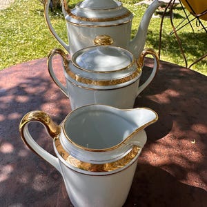 Puede incluir: Un juego de té de porcelana blanca con detalles dorados. El juego incluye una tetera, una azucarera y una jarra de crema. La tetera tiene tapa y boquilla curva. La azucarera tiene tapa y asas. La jarra de crema tiene boquilla y asa.