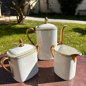 Könnte beinhalten: Weißes Porzellan-Teeservice mit goldenen Akzenten. Das Set beinhaltet eine Teekanne, eine Zuckerdose und einen Sahnekännchen. Jedes Teil hat einen goldenen Griff und Zierleisten mit einem grünen Blättermuster.