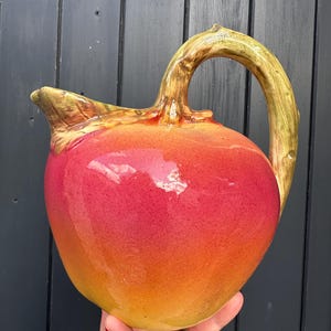 Può includere: Brocca in ceramica con una vivace sfumatura di rosso e arancione. Il manico e il beccuccio presentano un design strutturato a forma di ramo, con toni verdi e marroni. La brocca ha una finitura lucida e una forma organica unica.