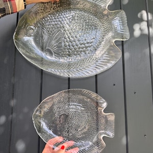 Op de afbeelding: Twee heldere, visvormige serveerschalen van glas. De schalen hebben gedetailleerde schubben en vinnen, met een textuur. Eén schaal wordt door een hand vastgehouden. De schalen worden gepresenteerd tegen een donkergrijze achtergrond.