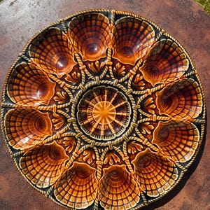 Könnte beinhalten: Eine Keramik-Austernservierplatte mit braunem und orangefarbenem Muscheldesign. Die Platte hat mehrere muschelförmige Fächer um ein zentrales Sonnenstrahl-Design mit Seil- und Blattdetails. Der Rand ist dunkelbraun.