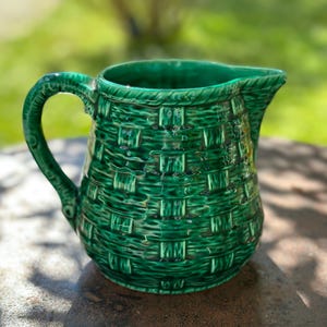 Può includere: Una brocca in ceramica verde con un motivo a intreccio di vimini. La brocca ha un manico e un beccuccio. La brocca è su una superficie di pietra con uno sfondo verde sfocato.