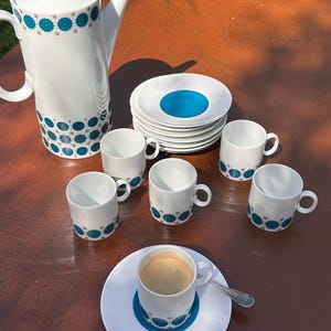 Könnte beinhalten: Weißes Keramik-Kaffeeservice mit türkisfarbenem Blumendesign. Das Set umfasst eine Kaffeekanne, Untertassen und sechs Kaffeetassen. Eine Tasse ist mit Kaffee gefüllt und steht auf einer Untertasse mit einem Löffel.