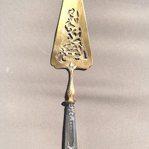 Peut inclure: Une pelle à gâteau vintage avec une lame triangulaire dorée ornée d'un motif ajouré. Le manche est argenté avec des détails floraux et un accent doré à la base de la lame. Un ustensile de service décoratif.