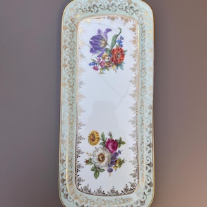 Op de afbeelding: Rechthoekige porseleinen serveerschaal met een lichtblauwe rand en gouden details. De schaal heeft bloemstukken in het midden en langs de randen. De bloemen omvatten paarse, rode, gele en witte bloemen.