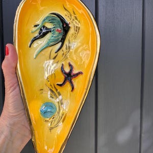 Könnte beinhalten: Eine dekorative, gelbe, muschelförmige Schale mit goldenem Rand. Die Schale zeigt einen schwarzen und teal Fisch, einen Seestern und eine blaue Muschel. Goldene Akzente und Details sind ebenfalls vorhanden.