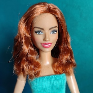 May include: A doll with auburn hair and a turquoise knitted top. The doll has a fair complexion, full lips, and detailed eye makeup. The doll's hair is styled in loose waves. The background is a solid teal color.