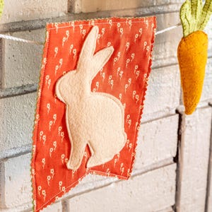 Könnte beinhalten: Ein festliches Osterbanner mit orangefarbenen Karotten aus Filz und roten Wimpeln mit weißen Hasensilhouetten. Das Banner ist an einer weißen Schnur befestigt und hängt vor einer weißen Backsteinmauer.