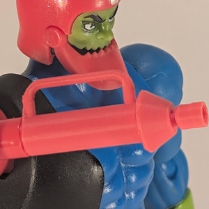 May include: A close-up of an action figure with a red helmet and weapon. The figure has a green face, blue torso, and black and green accents. The red weapon is held in the figure's hand.