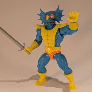 May include: A blue action figure with yellow armor and accents, holding a silver sword. The figure has a fish-like head with orange eyes and is posed in a dynamic stance, ready for action. The figure is approximately 6 inches tall.