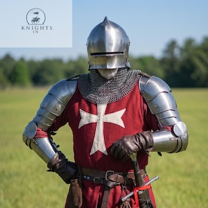 May include: A medieval knight in full armor stands in a grassy field. The knight wears a silver helmet, chainmail, and metal plating on the arms and legs. A red tunic with a white cross is visible. A sword hangs at the knight's side. The text "Knights In" is visible.