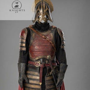 Puede incluir: Una armadura medieval completa, con casco, peto y protección para las piernas. La armadura es principalmente negra y roja con detalles dorados. Una espada está enfundada en la cintura, y una capa roja cae desde la cintura hacia abajo. Incluye el texto "Knights In".